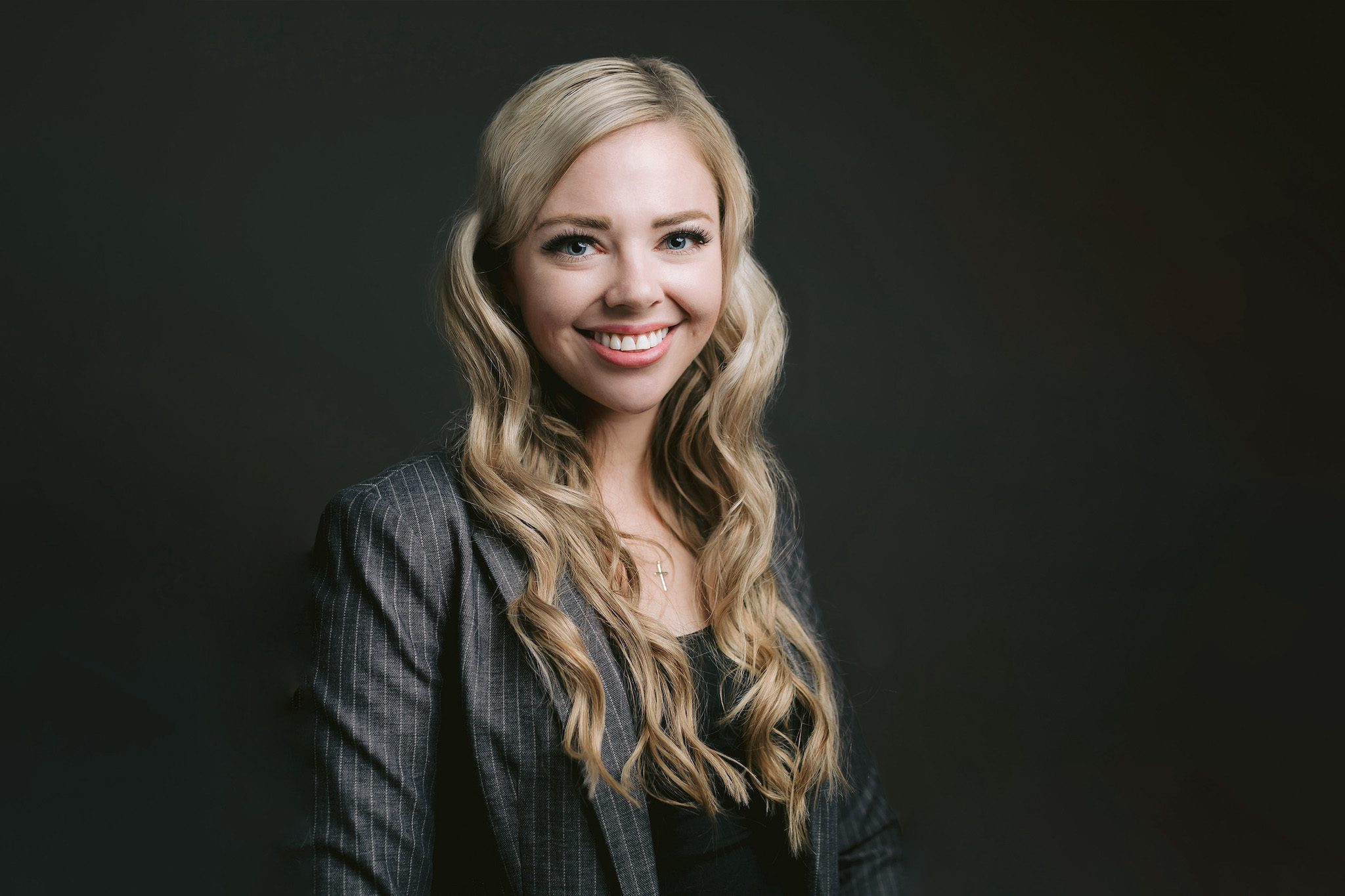 a blonde female professional in a business suit poses for a professional headshot, with a dark background highlighting her confidence with a beautiful smile.