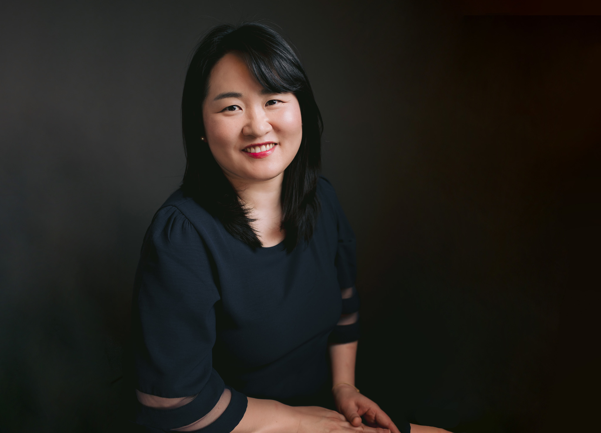 a woman in a black shirt sits on a chair, presenting a professional demeanor in her academic professor headshot.